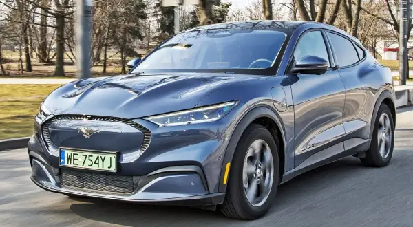 Niebieski Ford Mustang Mach-E jedzie po drodze, z widocznym logo konia na grillu.