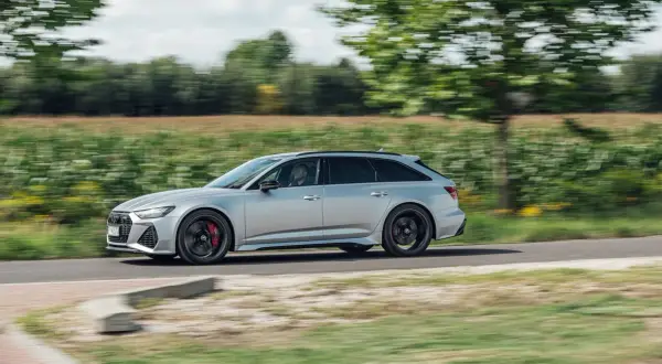 Srebrne Audi RS6 Avant pędzi po wiejskiej drodze, otoczone zielenią pól i drzew.