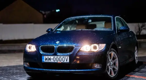 Ciemnoniebieskie bmw 325i z kroplami deszczu na karoserii, zaparkowane nocą na mokrym parkingu.