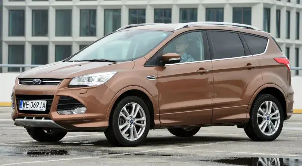 Brązowy Ford Kuga 2 z kierowcą w okularach i kratę, zaparkowany na mokrym parkingu przed nowoczesnym budynkiem.