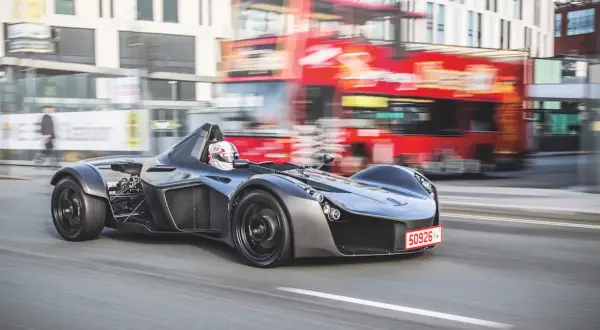 Niesamowity BAC Mono pędzi ulicą, mijając rozmazany czerwony autobus i budynki.