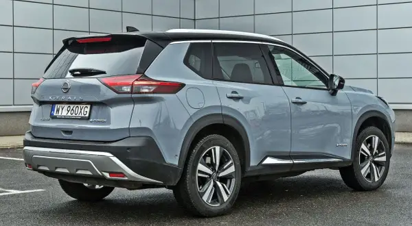 Szary Nissan X-Trail e-POWER z czarnym dachem i srebrnymi akcentami stoi na mokrym parkingu przed nowoczesnym budynkiem.