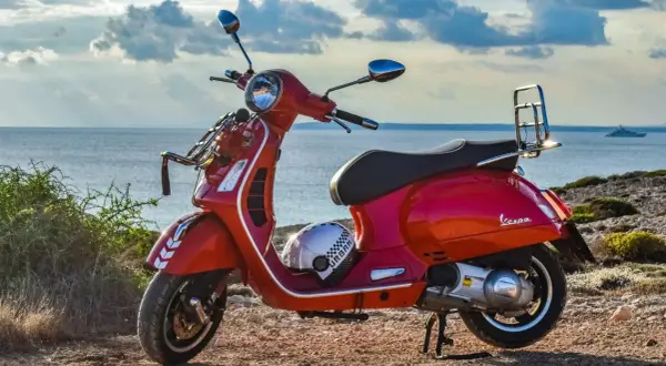 Czerwona Vespa na tle morza. Czy na skuter trzeba mieć prawo jazdy? Kask leży obok.