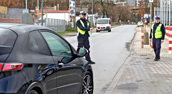 Policjant zatrzymuje czarne auto, być może kolejny raz, by wystawić mandat. W tle biały van i inni funkcjonariusze.