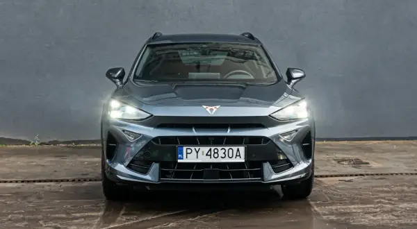 Szary Formentor Cupra z włączonymi światłami, zaparkowany przed betonową ścianą.