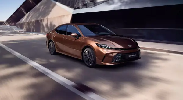 Brązowa Toyota Camry Hybrid 2024 pędzi po drodze, otoczona nowoczesną architekturą i cieniami drzew.