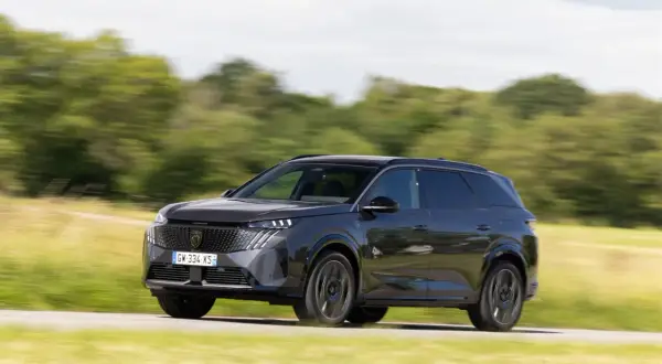 Ciemnoszary Peugeot 5008 jedzie po drodze wśród zielonych pól i drzew.