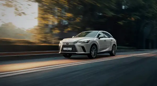 Biały Lexus RX 500h pędzi po drodze, otoczony rozmytymi drzewami i światłem zachodzącego słońca.