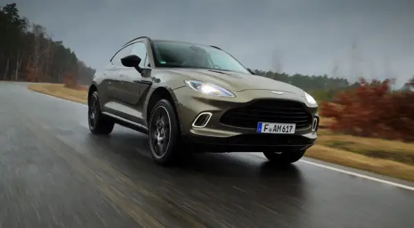 Elegancki Aston Martin SUV przemierza mokrą drogę wśród jesiennych drzew.