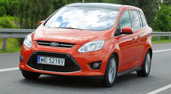 Pomarańczowy Ford C-Max 1.6 benzyna jedzie po drodze, odbijając chmury w szybie.