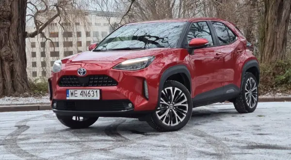 Czerwony Toyota Cross Hybrid na tle drzew i budynku. Auto stoi na ośnieżonej nawierzchni.