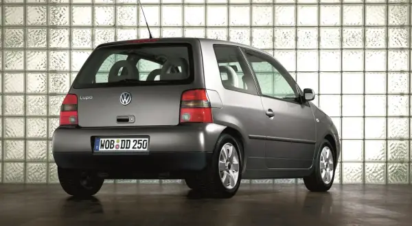 Szary Volkswagen Lupo stoi na ciemnej podłodze, na tle ściany z białych pustaków szklanych.