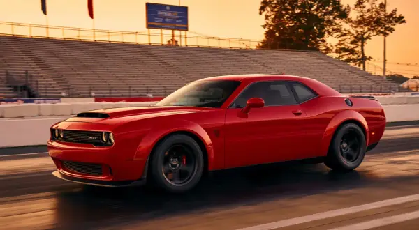 Czerwony Dodge Demon pędzi po torze wyścigowym podczas zachodu słońca, z trybunami w tle.