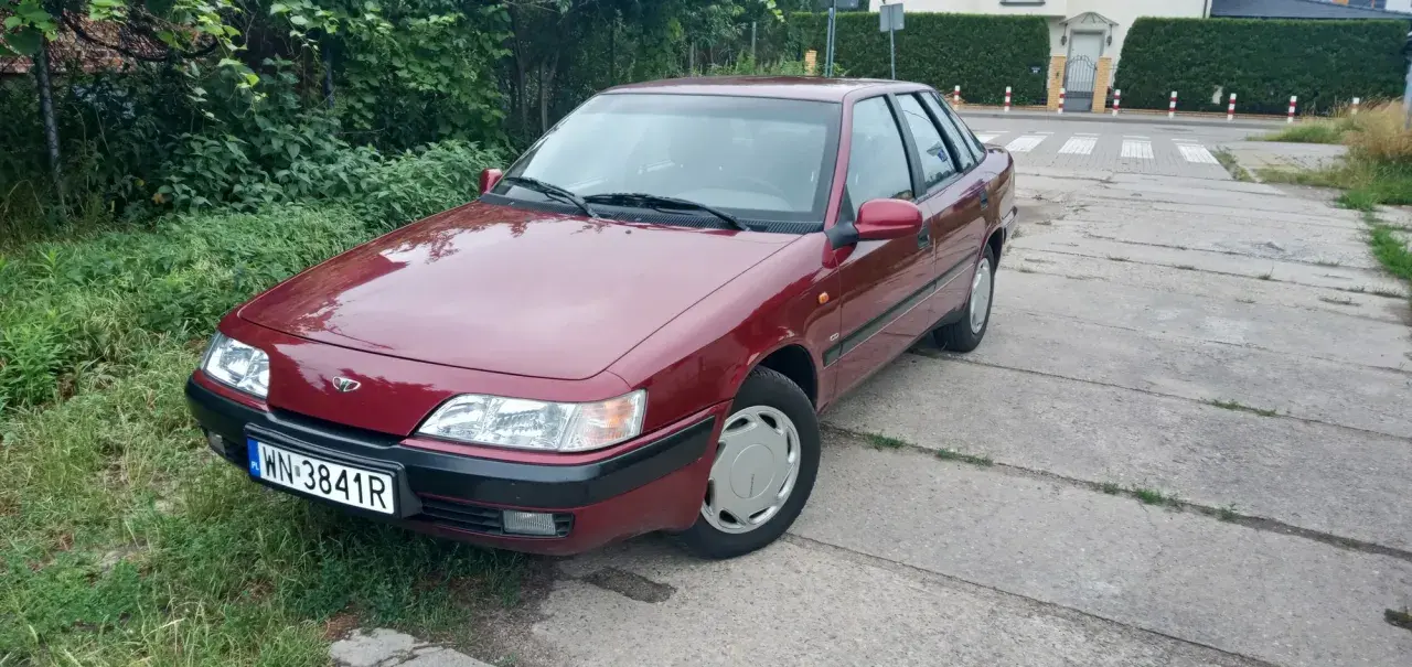 Czerwony Daewoo Espero z tablicą rejestracyjną WN-3841R zaparkowany przy zarośniętym poboczu i betonowej drodze.