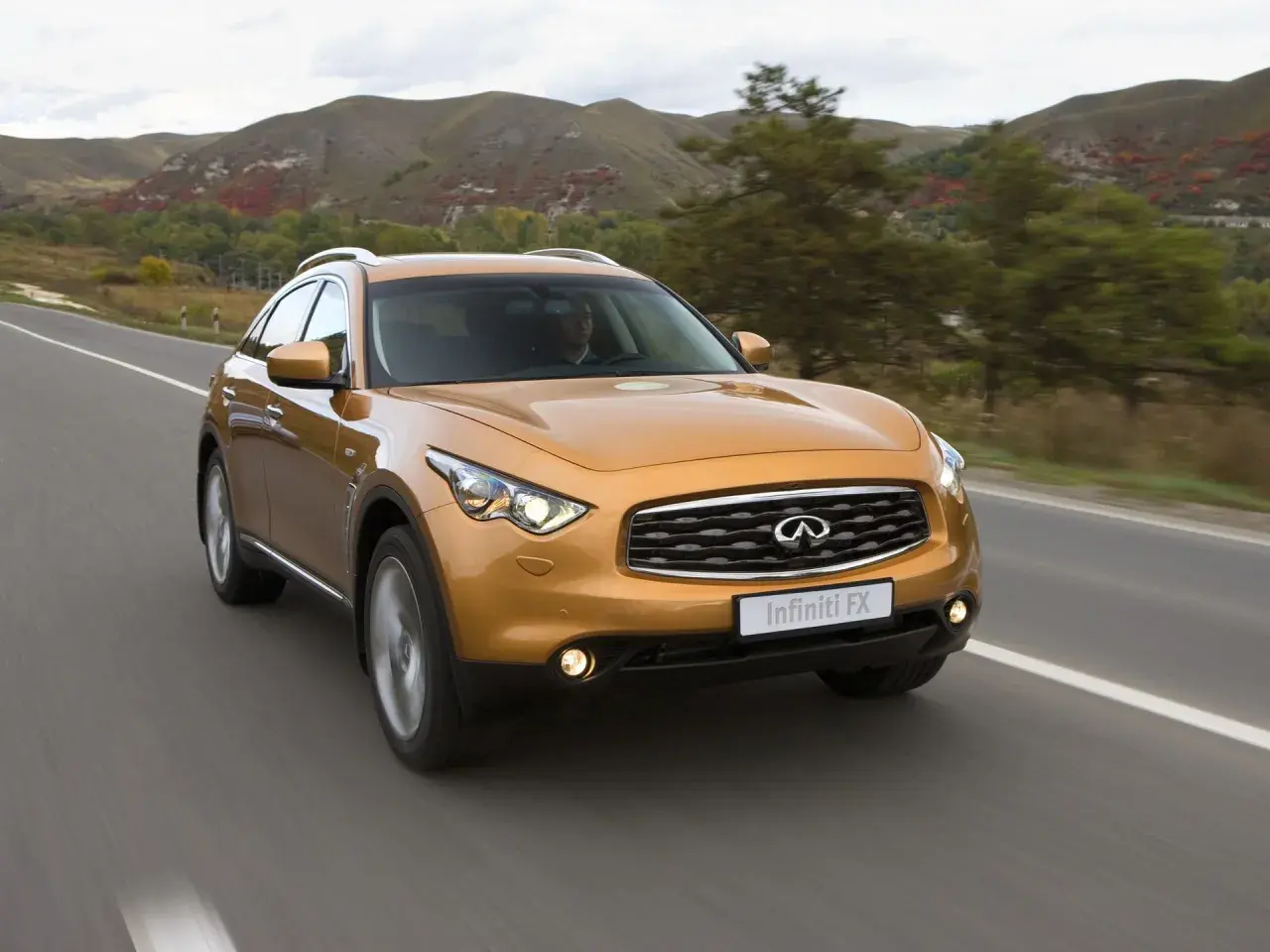Pomarańczowy Infiniti QX70 jedzie po drodze wśród jesiennych wzgórz.