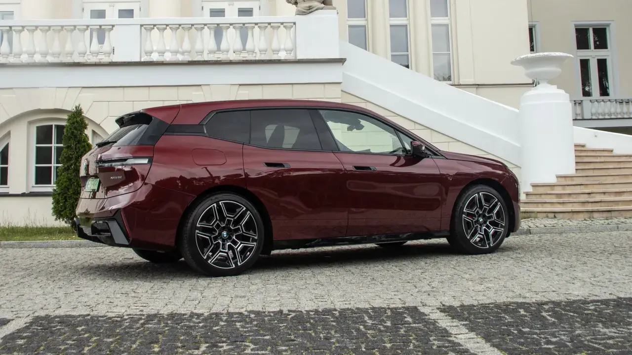 Bordowy BMW iX z eleganckimi felgami stoi przed luksusową posiadłością z kamienną nawierzchnią.