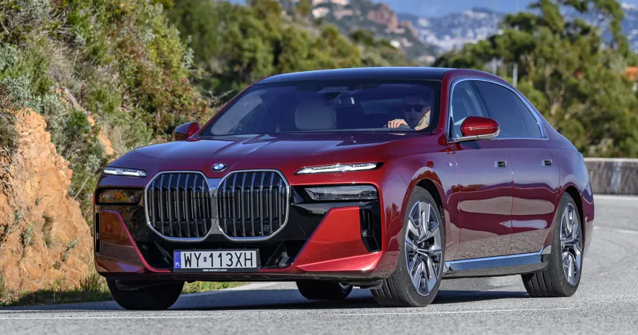 Czerwone BMW 740d z eleganckim czarnym dachem sunie po krętej drodze wśród zielonych wzgórz.