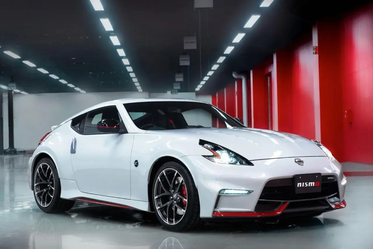 Biały Nissan 370Z Nismo z czerwonymi akcentami, lśniący w garażu z czerwonymi ścianami i rzędem świateł na suficie.