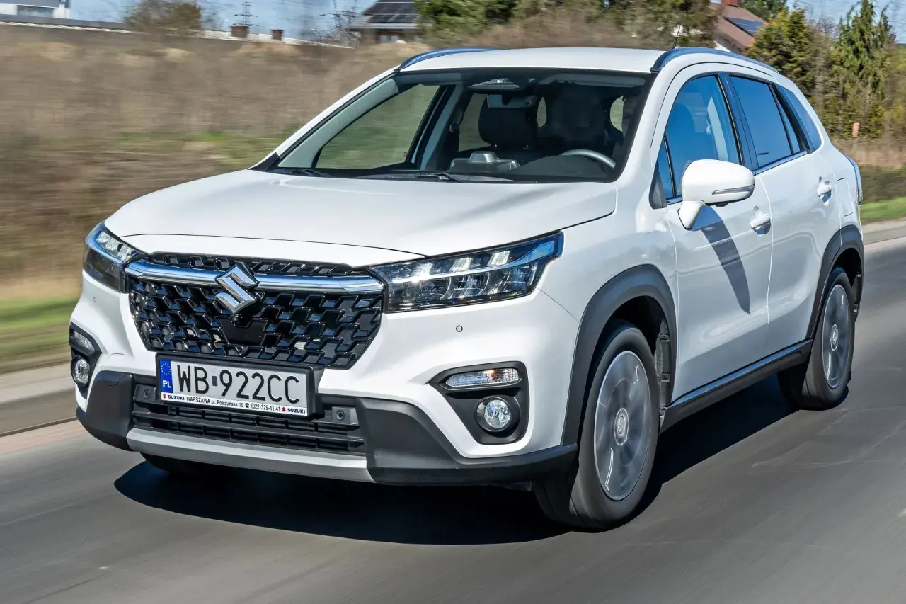 Biały Suzuki S-Cross jedzie po drodze, widoczny przód auta z grillem i światłami.