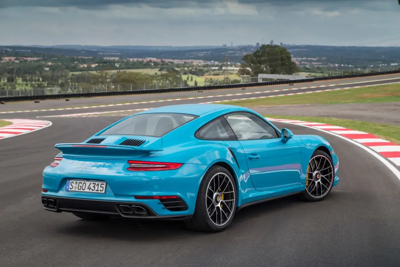 Błękitne Porsche 911 Turbo S na torze wyścigowym, z widokiem na miasto w tle.