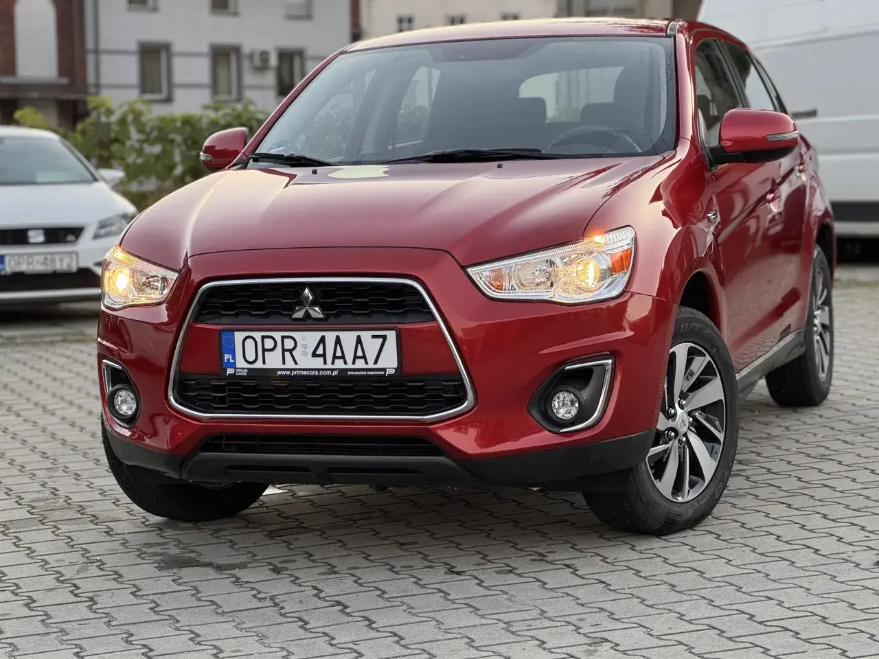 Czerwony Mitsubishi ASX 1.6 benzyna z tablicą rejestracyjną OPR 4AA7 zaparkowany na kostce brukowej.