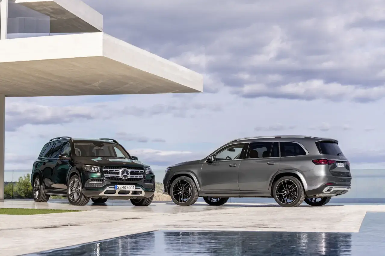 Dwa Mercedesy GLS 2025, jeden zielony, drugi szary, zaparkowane przed nowoczesnym budynkiem z basenem.