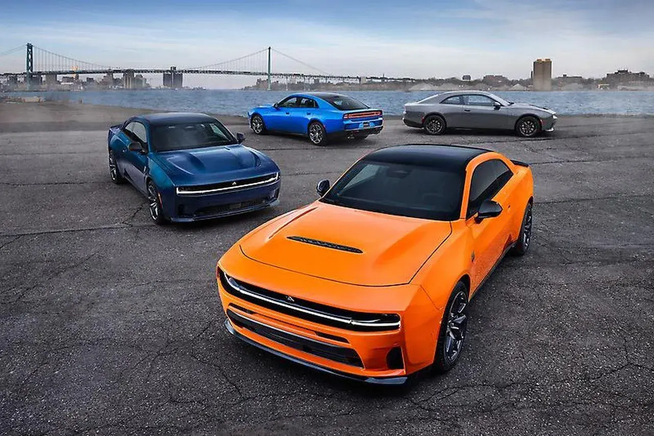 Pomarańczowy i niebieski Dodge Hellcat z innymi autami na tle mostu i miasta.