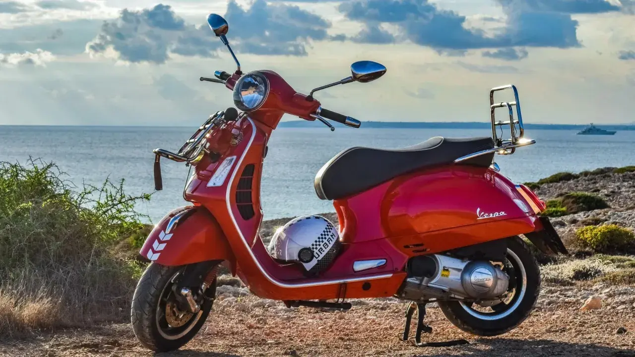 Czerwona Vespa na tle morza. Czy na skuter trzeba mieć prawo jazdy? Kask leży obok.