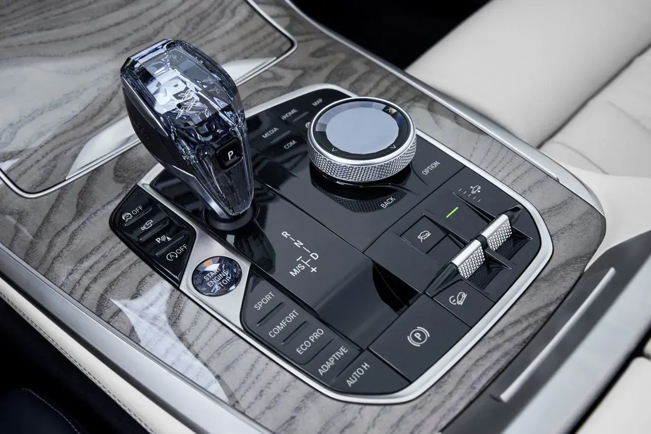 Kryształowa dźwignia zmiany biegów i pokrętło iDrive w BMW X7 2024, otoczone drewnianymi wstawkami i przyciskami trybów jazdy.