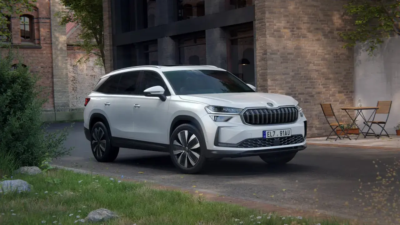 Biały Skoda Kodiaq 2025 zaparkowany przed ceglanym budynkiem, z zielenią i stolikiem z krzesłami w tle.