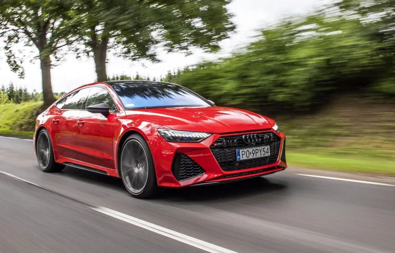 Czerwone Audi RS 7 pędzi po drodze, otoczone zielenią drzew.