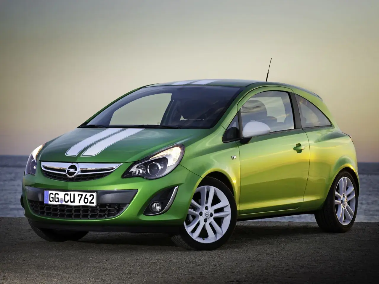 Zielony Opel Corsa D 1.2 benzyna z białymi pasami na masce, zaparkowany przy plaży.