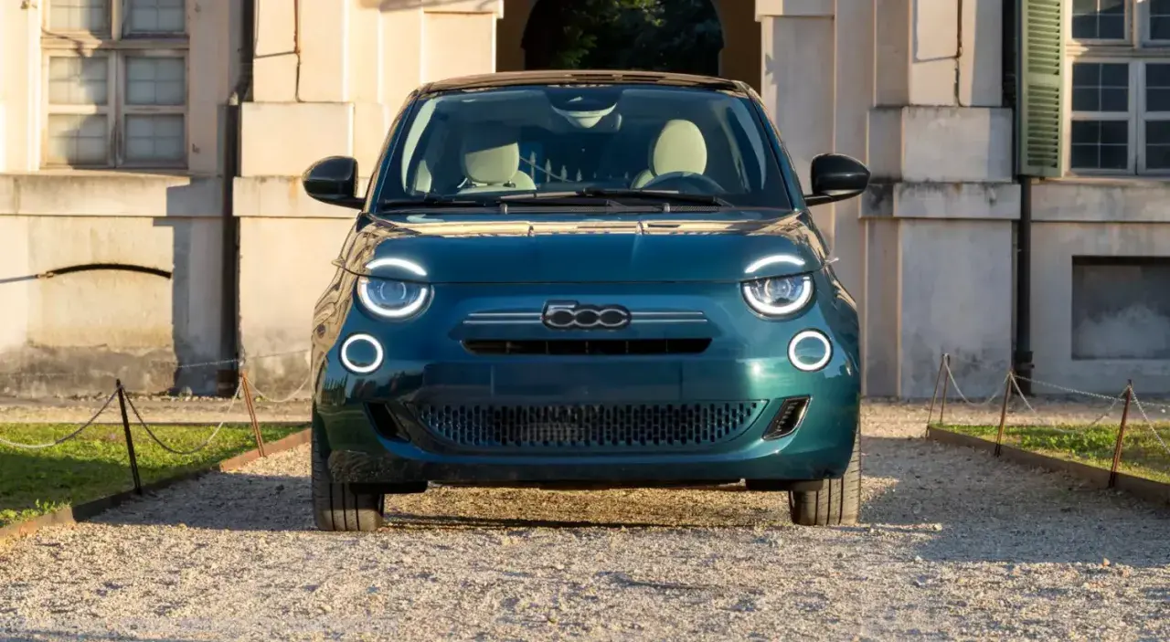 Nowy Fiat 500 w kolorze głębokiej zieleni, z charakterystycznymi okrągłymi światłami, zaparkowany przed zabytkowym budynkiem. Sprawdź aktualną fiat 500 cena!