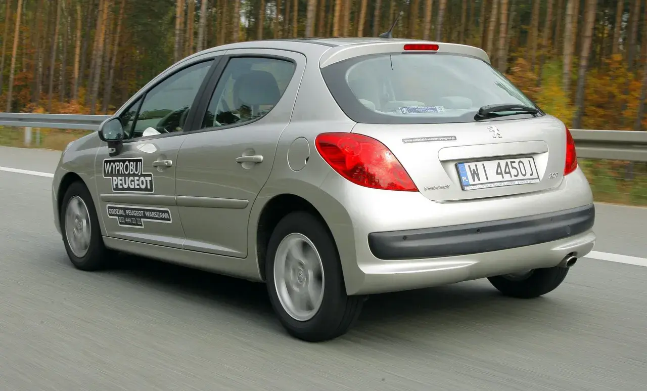 Srebrny Peugeot 207 1.4 benzyna w ruchu drogowym, z naklejkami "Wypróbuj Peugeot" i danymi oddziału.