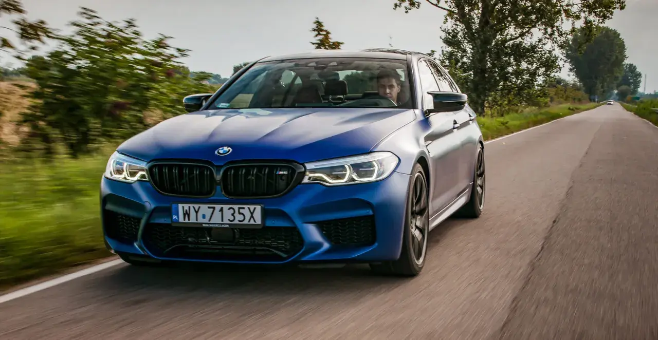 Niebieskie BMW M5 F90 pędzi po wiejskiej drodze, kierowca skupiony na trasie.