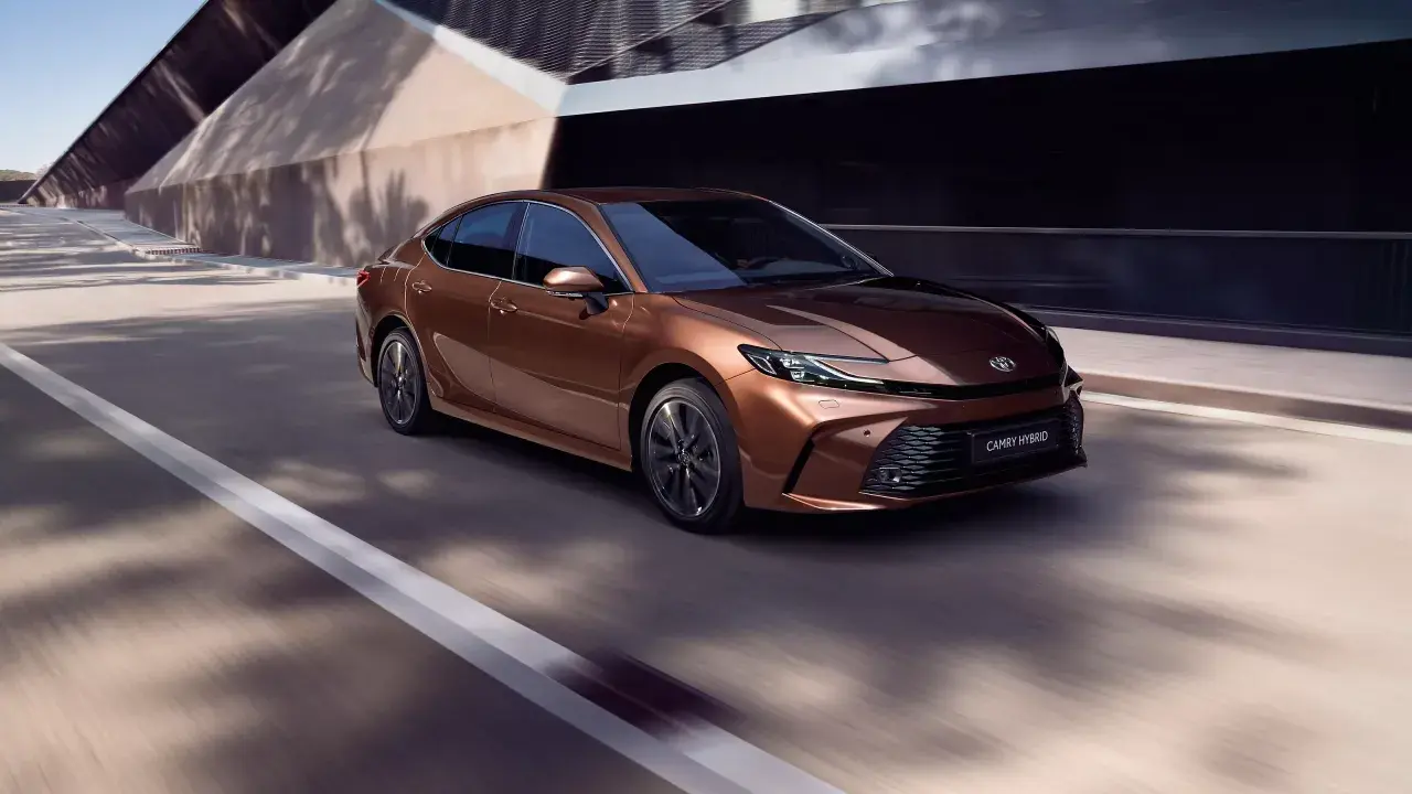 Brązowa Toyota Camry Hybrid 2024 pędzi po drodze, otoczona nowoczesną architekturą i cieniami drzew.