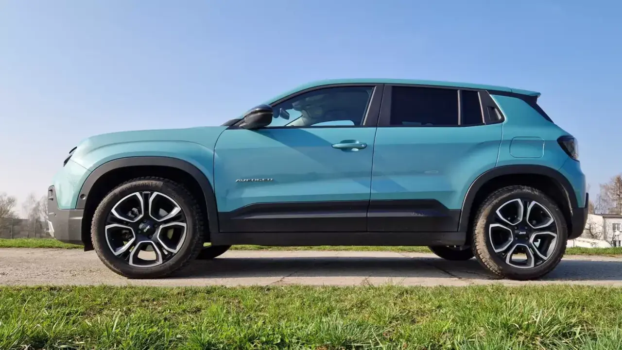 Turkusowy Jeep Avenger, marka samochodu na wprost, stoi na tle błękitnego nieba i zielonej trawy.