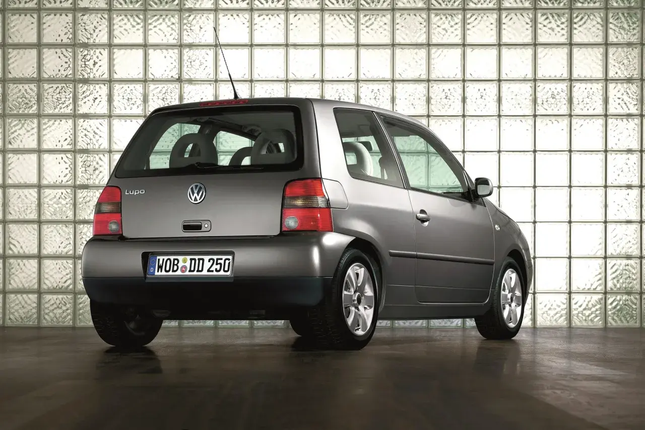 Szary Volkswagen Lupo stoi na ciemnej podłodze, na tle ściany z białych pustaków szklanych.