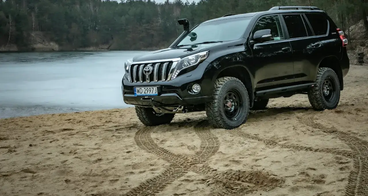 Czarny Toyota Land Cruiser 2024 na piaszczystym brzegu jeziora, gotowy na przygodę.