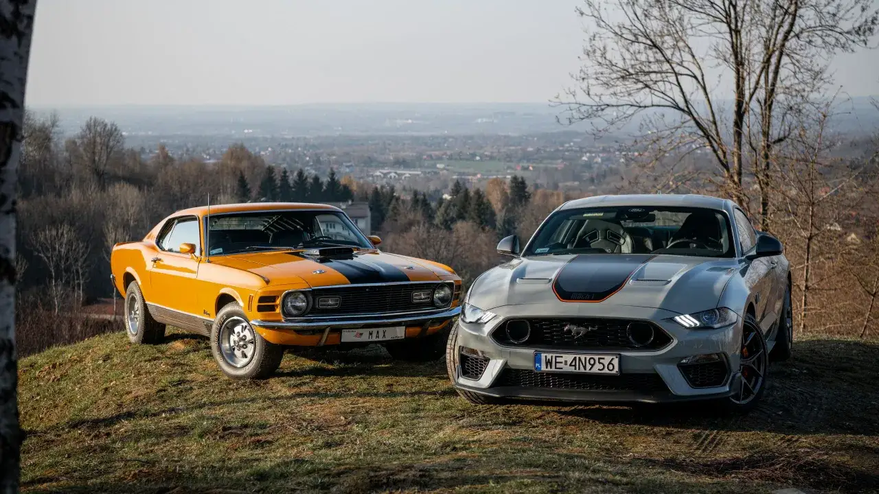 Dwa Fordy Mustang: klasyczny pomarańczowy z lat 70. i nowoczesny szary, oba w wersji Mustang Mach 1, stoją na wzgórzu z widokiem na dolinę.