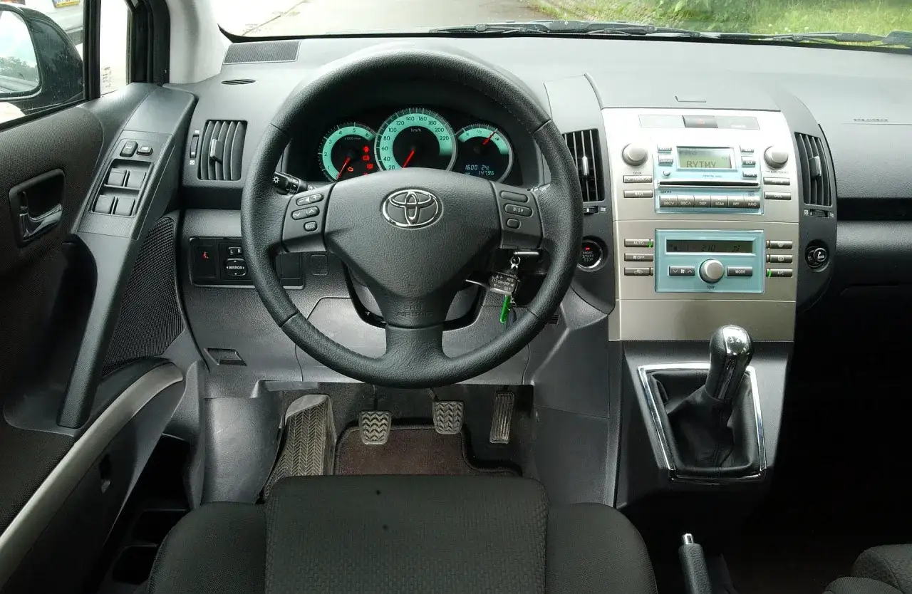 Wnętrze Toyoty Verso 1.8 benzyna. Widok na deskę rozdzielczą, kierownicę z logiem Toyoty, panel radia i dźwignię zmiany biegów.