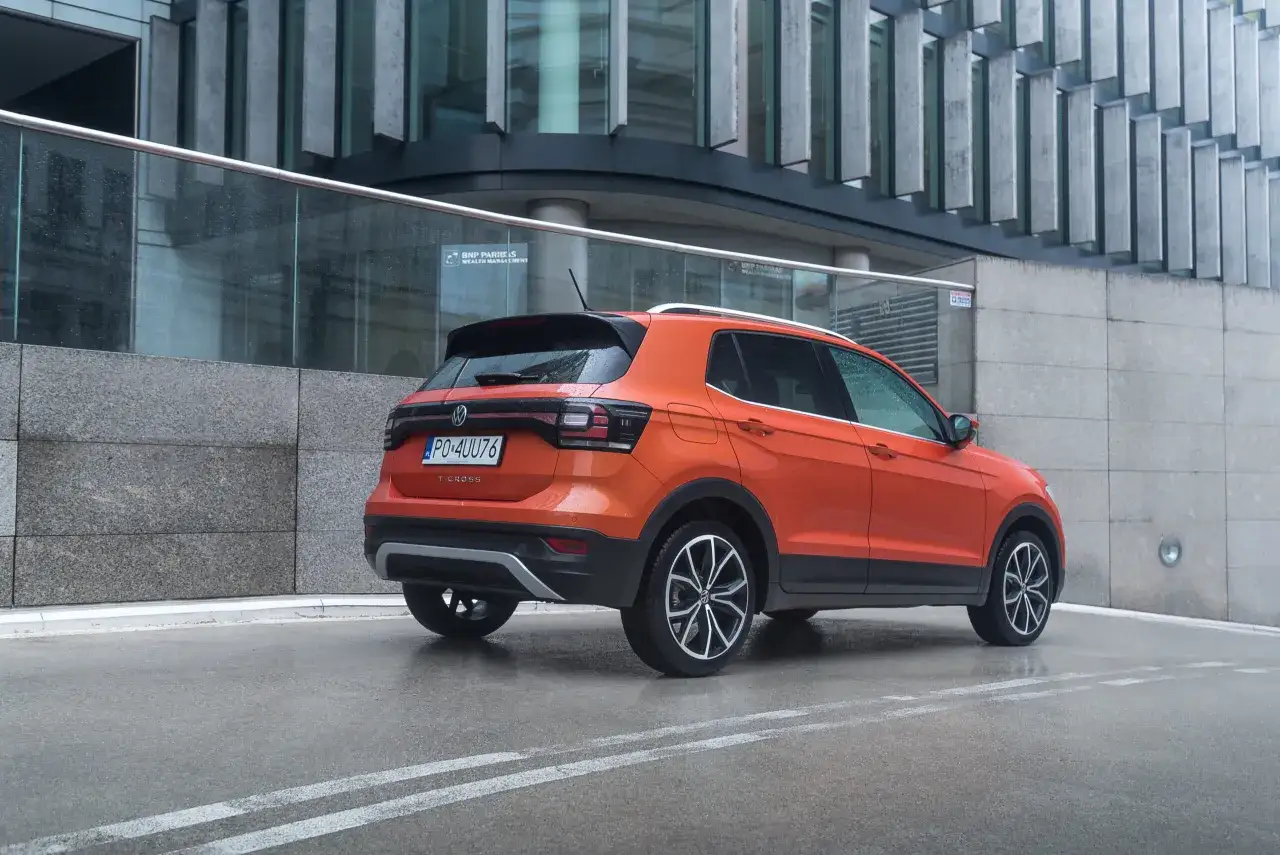 Pomarańczowy Volkswagen T-Cross stoi na mokrej nawierzchni przed nowoczesnym budynkiem.