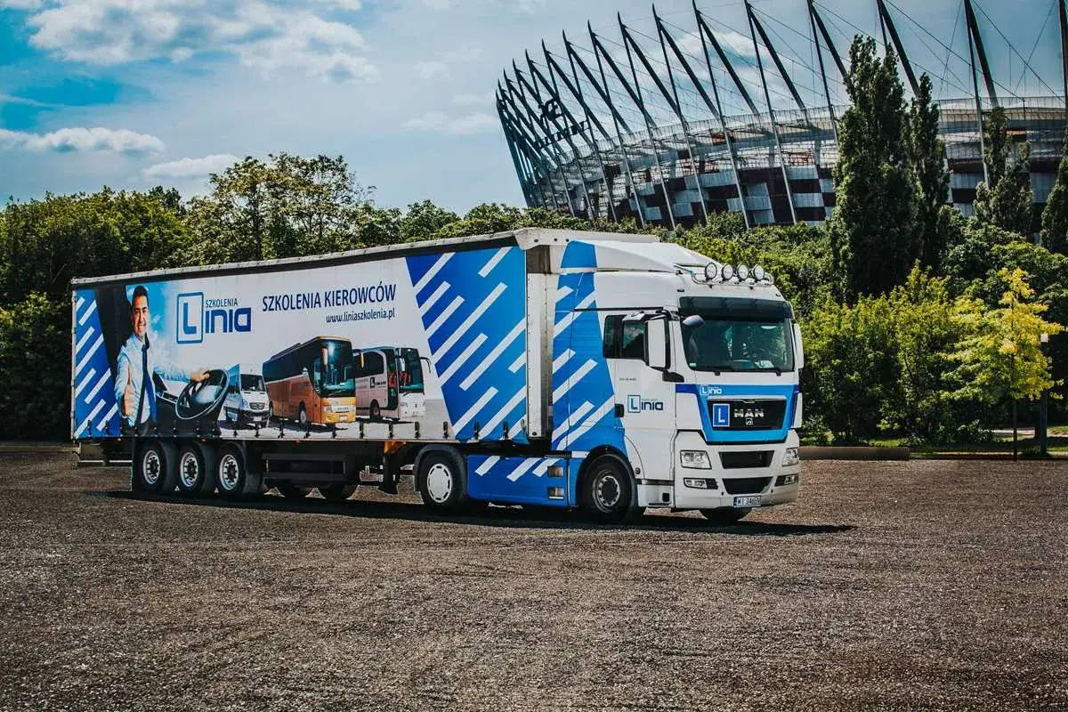 Ciężarówka z reklamą szkoleń kierowców, w tym prawo jazdy kat C+E. Na naczepie widnieje mężczyzna z kierownicą i grafiki pojazdów.