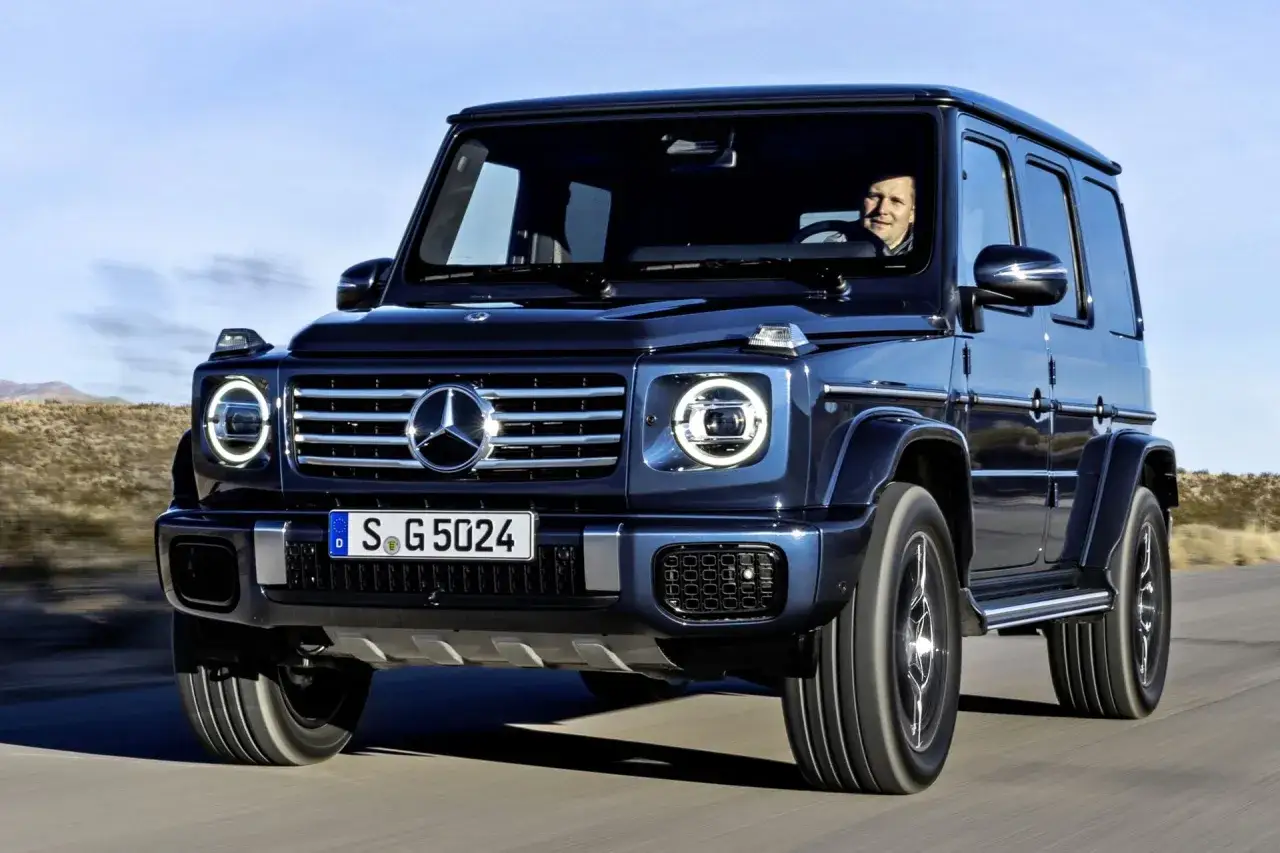 Ciemnoniebieski Mercedes G Klasa jedzie po drodze. Kierowca uśmiecha się. Sprawdź aktualną mercedes g klasa cena.
