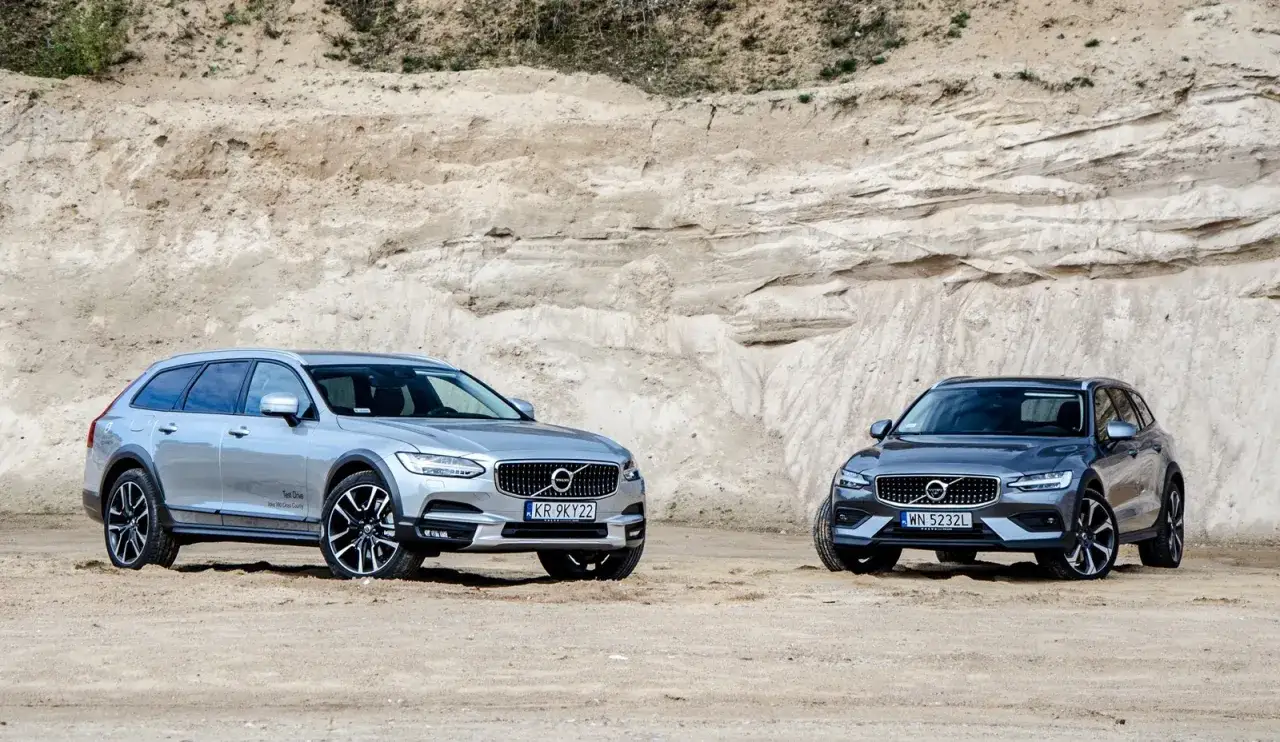 Dwa samochody Volvo Cross Country, srebrny i grafitowy, stoją na piaszczystym podłożu przed skalistym zboczem.