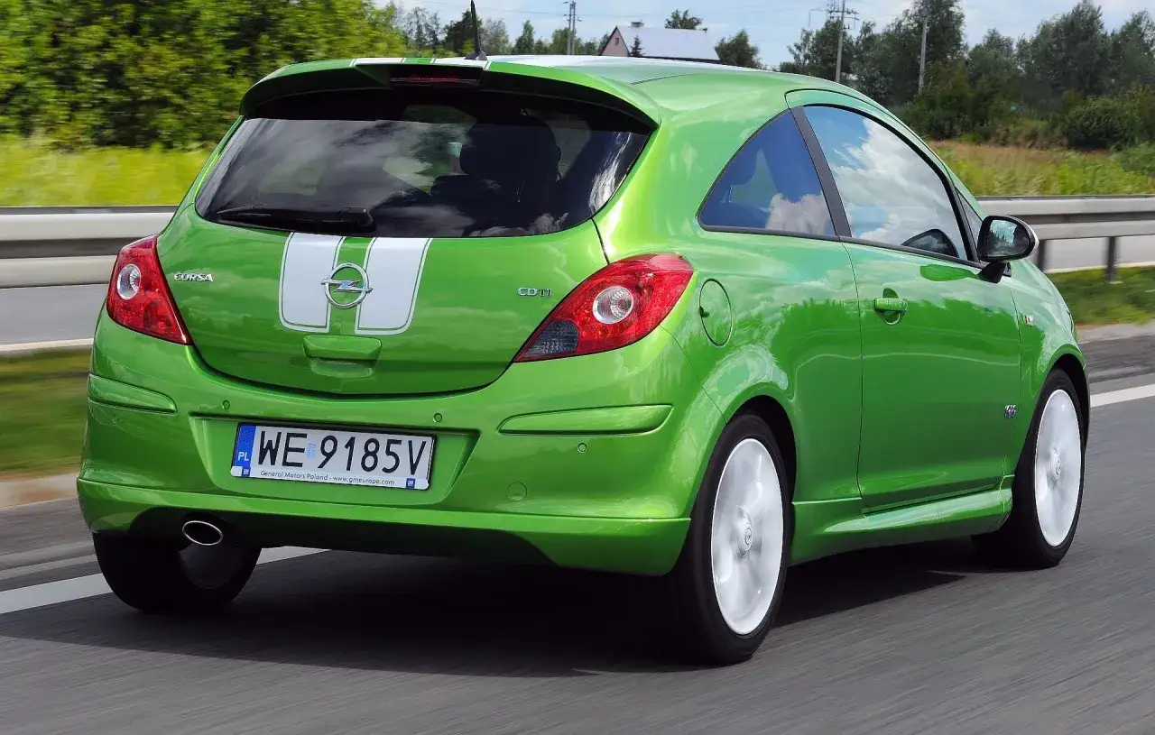 Zielony Opel Corsa D 1.2 benzyna z białymi pasami na klapie bagażnika jedzie po drodze.