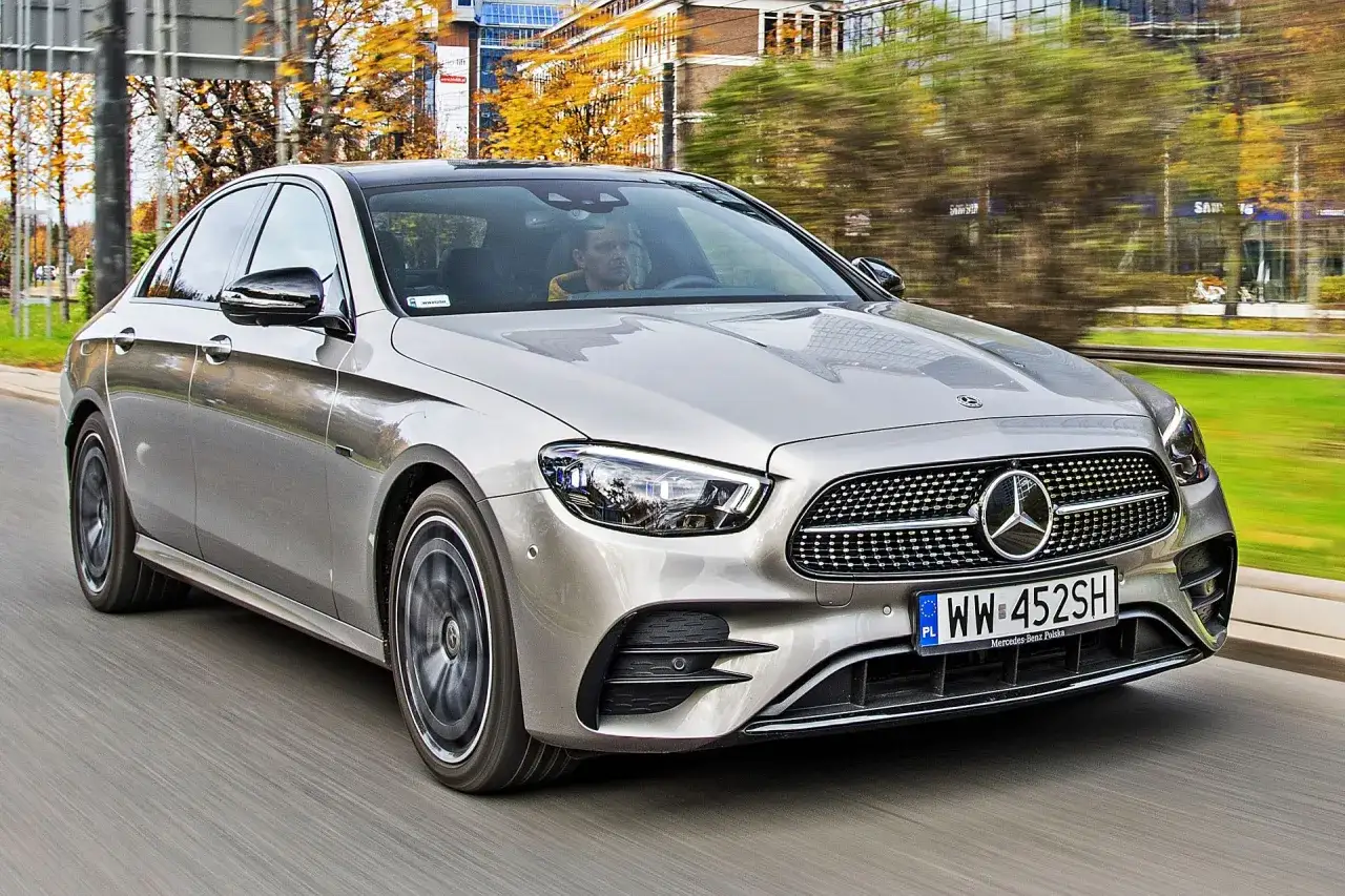 Srebrny Mercedes E220d w ruchu miejskim, z kierowcą za kierownicą. Jesienne drzewa i budynki w tle.