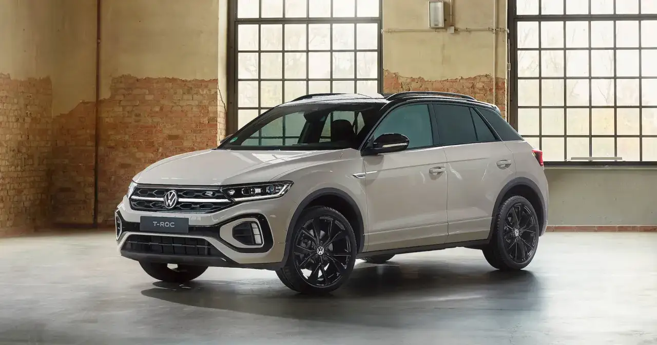 Nowy Volkswagen T-Roc 2025 w kolorze piaskowym, z czarnymi felgami i dachem, prezentuje się stylowo w industrialnym wnętrzu.