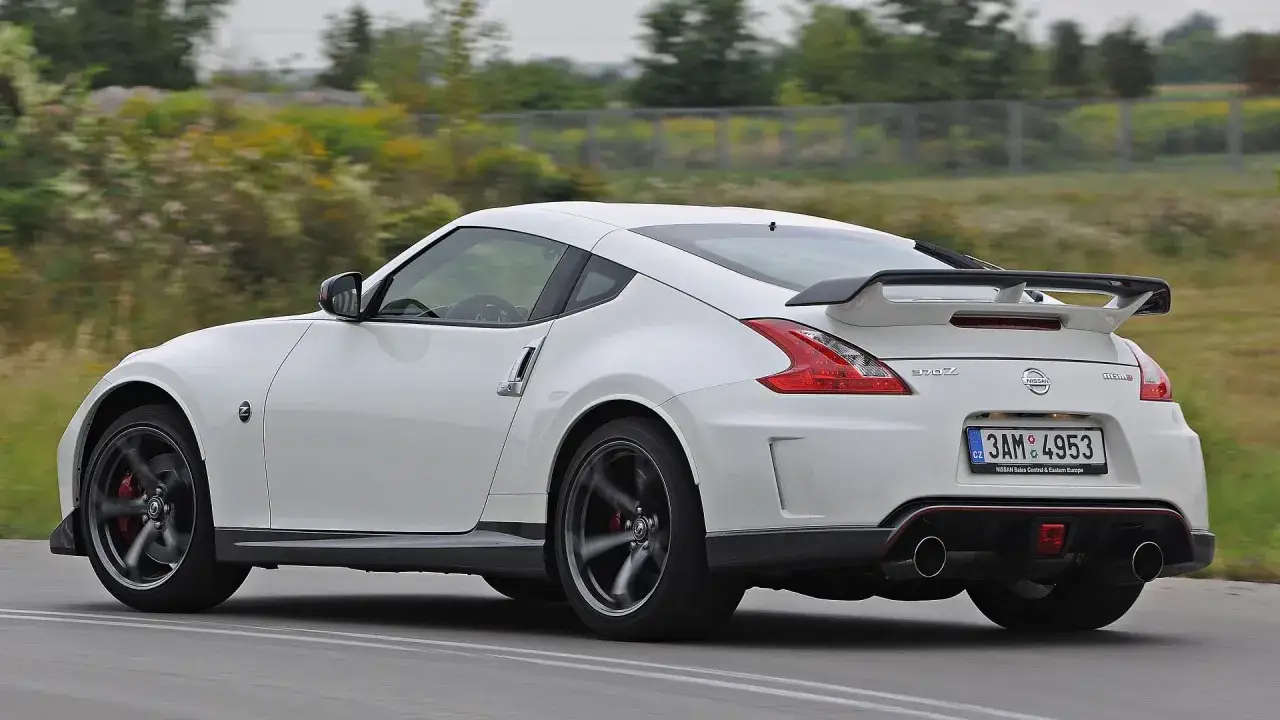 Biały Nissan 370Z Nismo z charakterystycznym tylnym spojlerem i dyfuzorem pędzi po drodze.
