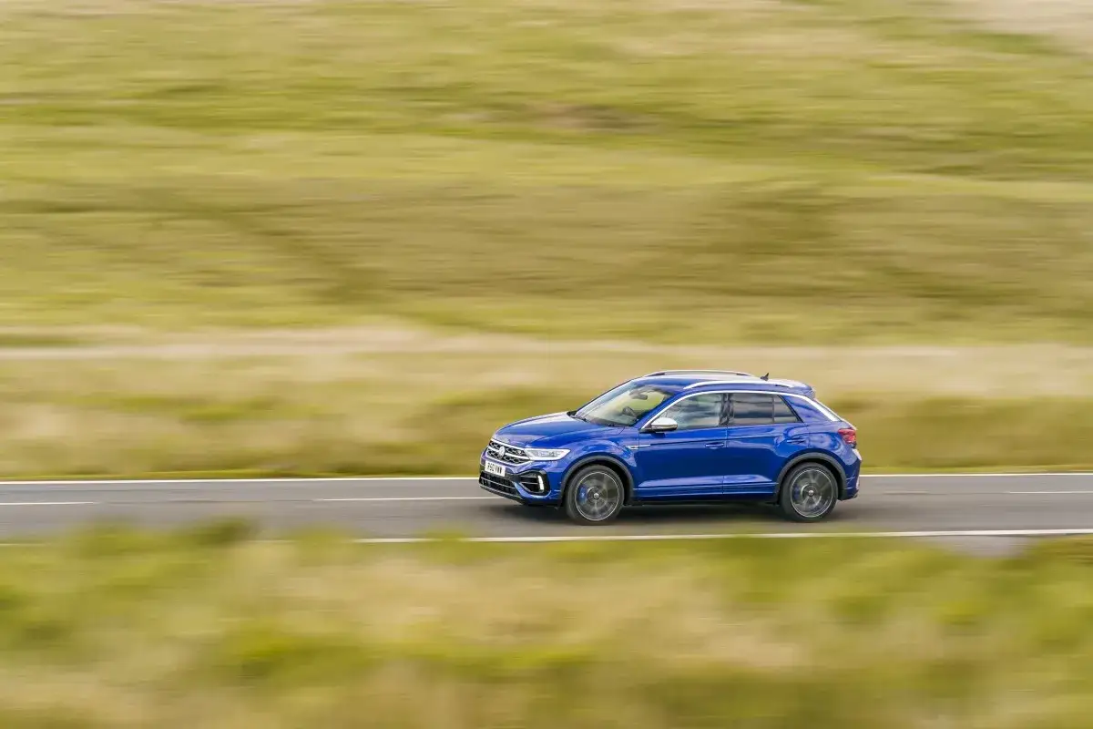 Niebieski Volkswagen T-Roc pędzi po drodze, a krajobraz rozmywa się w tle. Czy to początek przygody, czy może podróż do mechanika z powodu usterki?